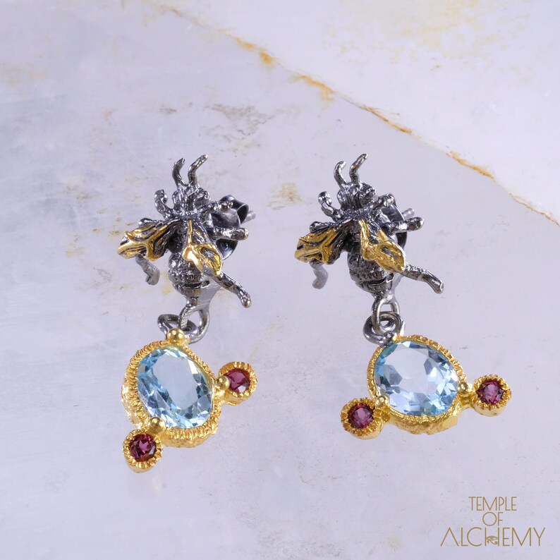 May include: A pair of silver and gold earrings featuring a bee design with a dangling blue gemstone and red accents. The earrings are a statement piece with a unique and intricate design.