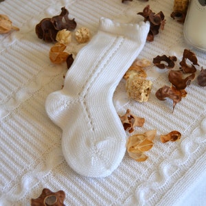 May include: A white knitted baby sock with a lacy pattern. The sock is lying on a white crocheted blanket with dried flower petals.