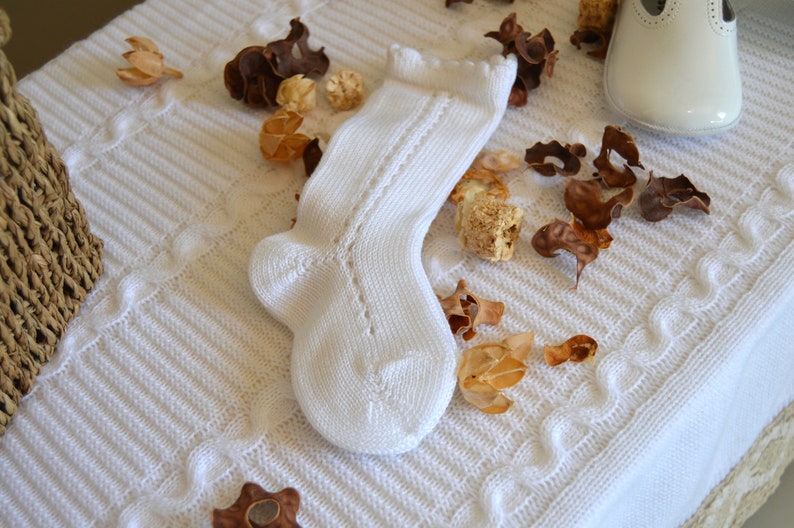 May include: A white knitted baby sock with a lacy pattern. The sock is lying on a white crocheted blanket with dried flower petals.