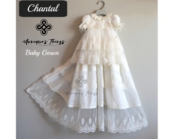 Può includere: Un abito bianco per neonato con dettagli in pizzo e un colletto a balze. L'abito ha più strati di tulle e pizzo, creando un look fluido ed elegante. L'abito è appeso a un appendiabiti.