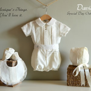 May include: A white baby outfit with a white bib, hat, and shoes. The outfit has a white lace trim and a white satin ribbon. The text "Monique's Things. You'll love it." is visible in the image.