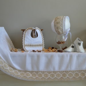 May include: A white baby blanket with a beige lace trim, a white bib with beige lace trim, a white bonnet with beige lace trim, and a pair of white baby shoes.