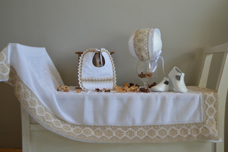 May include: A white baby blanket with a beige lace trim, a white bib with beige lace trim, a white bonnet with beige lace trim, and a pair of white baby shoes.