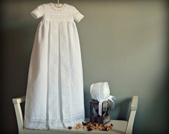 May include: A white christening gown with lace trim and a matching bonnet. The gown is hanging on a hanger and the bonnet is sitting on a wooden stand.