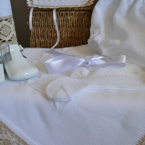 May include: White baby shoes, socks, and a blanket with lace trim. The items are arranged on a white surface with a wicker basket in the background.