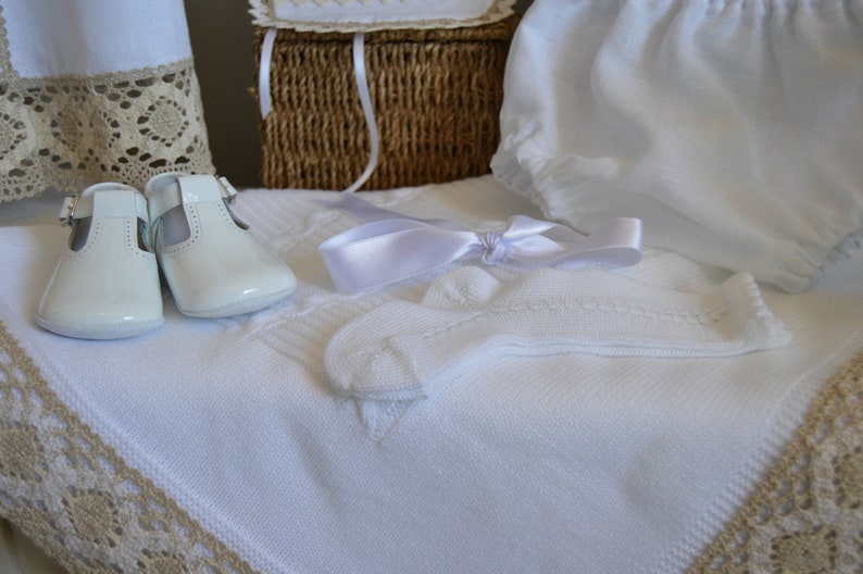 May include: White baby shoes, socks, and a blanket with lace trim. The items are arranged on a white surface with a wicker basket in the background.