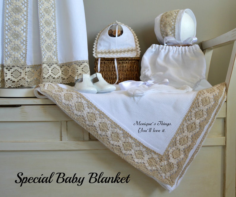 May include: A white baby blanket with a beige crocheted border. The blanket is folded and displayed on a white wooden bench with a small white bonnet, white diaper, and white shoes. The text "Monique's Things, You'll love it." is embroidered on the blanket.