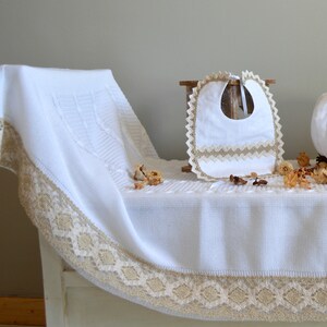 May include: White knitted baby blanket with a lace trim and a white bib with a lace trim. The blanket and bib are on a wooden surface.