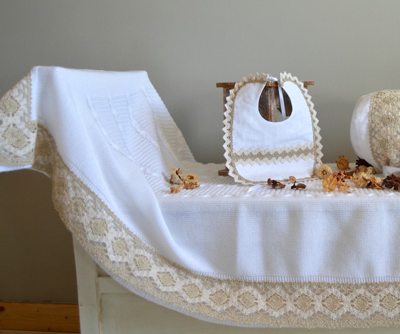 May include: White knitted baby blanket with a lace trim and a white bib with a lace trim. The blanket and bib are on a wooden surface.