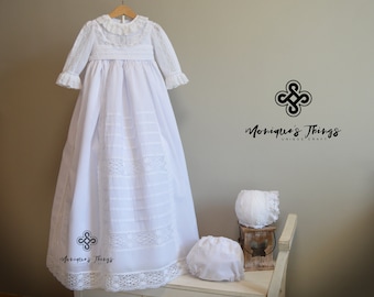 May include: White christening gown with lace trim and a matching bonnet. The gown has a ruffled collar and long sleeves. The gown is hanging on a wooden hanger. The text "Monique's Things Unique Craft" is visible in the image.