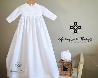May include: White christening gown with long sleeves and embroidered details. The gown is hanging on a wooden hanger and is displayed on a white dresser. The text "Monique's Things Unique Craft" is visible in the image.