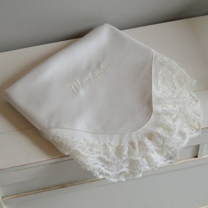 以下が含まれることがあります： レースの縁取りと「マテア」という名前が刺繍された白いベビーブランケットです。