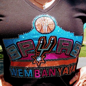 May include: A black t-shirt with a glittery design featuring a basketball, the San Antonio Spurs logo, and the text "Wembanyama".