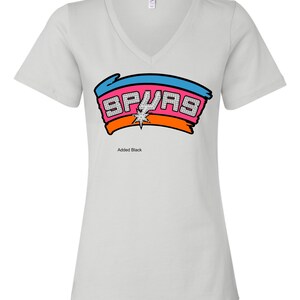 May include: A light gray V-neck t-shirt featuring the word "SPURS" in a colorful design. The design includes a blue and orange banner with the word "SPURS" in a pink, diamond-like font, and a star in the center. The text "Added Black" is below the design.