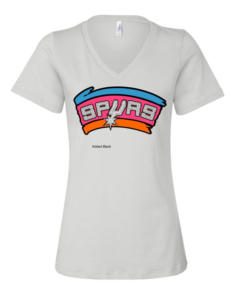 May include: A light gray V-neck t-shirt featuring the word "SPURS" in a colorful design. The design includes a blue and orange banner with the word "SPURS" in a pink, diamond-like font, and a star in the center. The text "Added Black" is below the design.