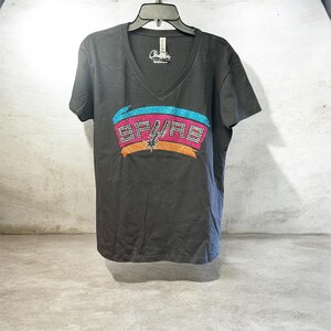 May include: A dark gray V-neck t-shirt featuring a colorful graphic of the San Antonio Spurs logo. The logo includes the team name and a spur design, with accents of pink, blue, orange, and silver. The shirt is hanging on a black hanger.