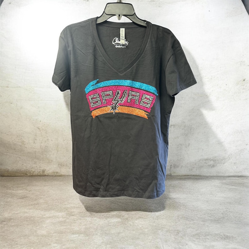 May include: A dark gray V-neck t-shirt featuring a colorful graphic of the San Antonio Spurs logo. The logo includes the team name and a spur design, with accents of pink, blue, orange, and silver. The shirt is hanging on a black hanger.