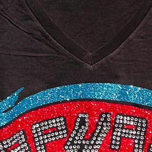 May include: Dark gray V-neck t-shirt with a glittery design. The design features the word "BEACH" in silver rhinestones, set against a red and orange background, with a blue glitter arc above. The shirt has a tag at the neckline.