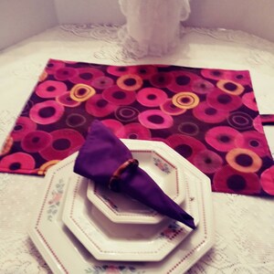 May include: A table setting featuring a stack of white octagonal plates with floral designs, a folded purple napkin with a wooden ring, and a patterned placemat with pink and yellow circles. A decorative white object is in the background.
