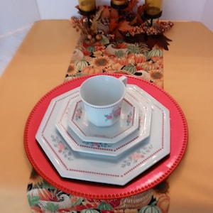 May include: A red octagonal charger plate with a white octagonal dinner plate, a white saucer, and a white teacup with pink floral designs. The plates and cup are set on a table runner with a fall-themed pattern of pumpkins, sunflowers, and turkeys.
