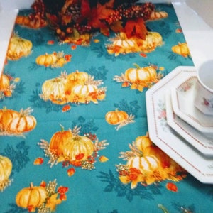 Op de afbeelding: Een turquoise tafelloper met een herhaald patroon van oranje pompoenen en herfstbladeren. De loper is versierd met een middelpunt van kaarsen, bladeren en bessen. Witte borden en een theekopje rechts.