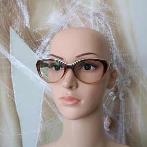 May include: A brown and gold cat-eye style eyeglasses frame with clear lenses. The frame has a slight upward curve at the brow line.