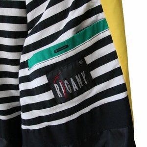 May include: A black and white striped jacket with a green and black pocket and a yellow panel. The jacket has a label that says "RIGANY" in red.