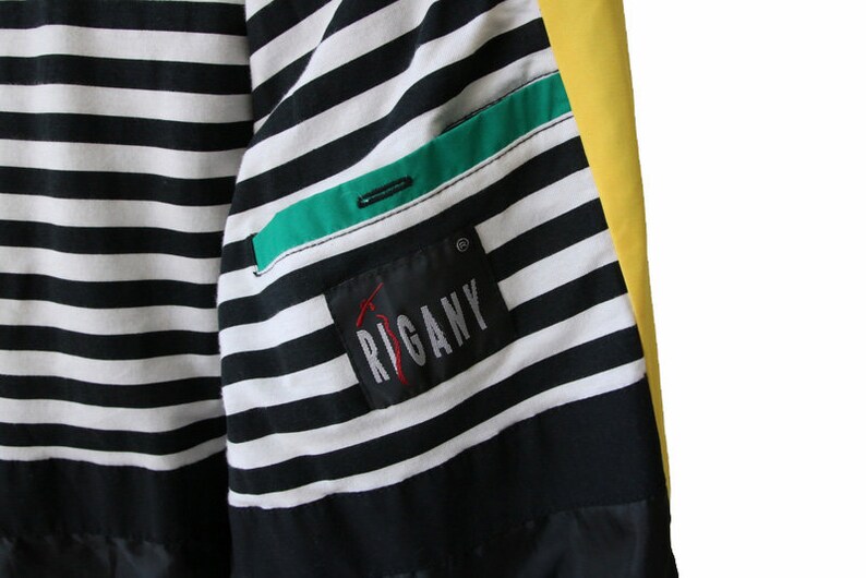 May include: A black and white striped jacket with a green and black pocket and a yellow panel. The jacket has a label that says "RIGANY" in red.