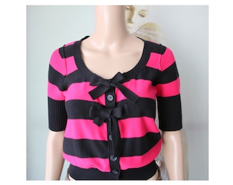 Op de afbeelding: Een zwart-roze gestreepte cardigan met knoopsluiting en strikken aan de voorkant.