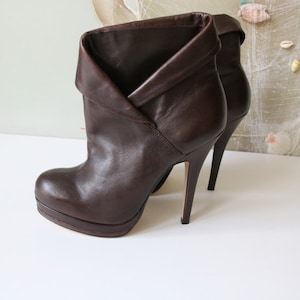 May include: A pair of brown leather ankle boots with a high heel and a folded-over cuff. The boots are on a white surface.