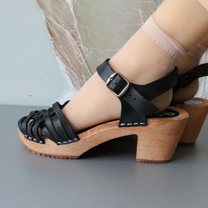 Może przedstawiać: Czarne skórzane sandały z plecionym paskiem na palcach i paskiem na kostce ze złotą klamrą. Sandały mają drewnianą podeszwę i obcas. Wysokość obcasa wynosi około 7.6 cm.