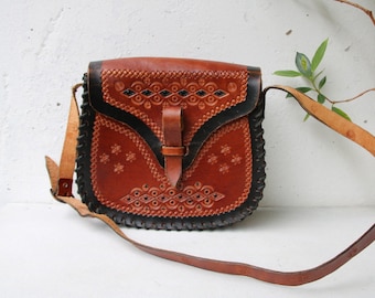 May include: A brown leather crossbody bag with intricate embossed floral and geometric patterns. The bag features a black border and a long, adjustable strap. A leather flap with a buckle closure secures the bag's contents.