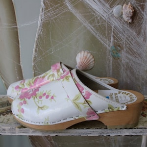 May include: A pair of white clogs with a floral print featuring pink roses and green leaves. The clogs have a wooden sole and a small heel. The clogs are sitting on a white shelf with a floral pattern.