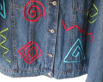 以下が含まれることがあります： カラフルな刺繍の幾何学模様が施された青いデニムジャケット。模様には、渦巻き、正方形、三角形、波線などがあります。