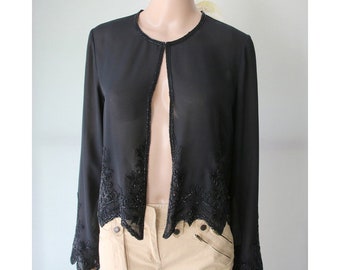 May include: A black open-front jacket with long sleeves. The jacket features intricate beaded embellishments along the edges and cuffs. The neckline is also trimmed with beading. The jacket is displayed on a mannequin.