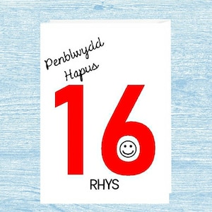 May include: A white card with the text "Penblwydd Hapus" in black. The number 16 is in red with a smiley face in the center of the 6. The name "Rhys" is written in black below the number.