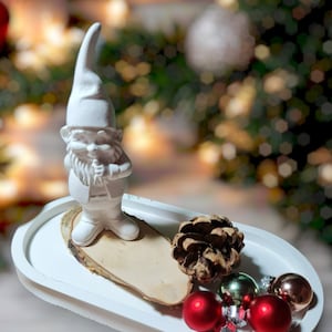 Peut inclure: Une figurine de gnome en céramique blanche debout sur une tranche de bois avec des pommes de pin et des ornements rouges et dorés dans un plateau ovale blanc.