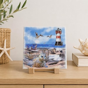 Könnte beinhalten: Eine quadratische Keramikfliese mit einer Aquarellillustration von zwei Robben, die an einem Sandstrand ruhen. Die Szene zeigt einen rot-weiß gestreiften Leuchtturm, blaue Meereswellen, zwei fliegende Möwen und zwei blaue Strandstühle.