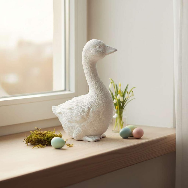Unbemalte Gans auf heller Fensterbank in ruhiger Frühlings-/Osterszene mit zarter Vase und Naturdeko