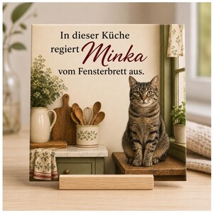 Peut inclure: Une plaque décorative carrée représentant un chat tigré brun assis sur un rebord de fenêtre. Le texte indique, In dieser Küche regiert Minka vom Fensterbrett aus. L'image montre une cuisine avec un pichet en céramique et des cuillères en bois.
