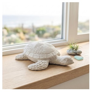 Op de afbeelding: Een gedetailleerd, crèmekleurig keramiek schildpadbeeldje met een getextureerd schild en vinnen. De schildpad staat op een houten vensterbank, naast een kleine luchtplant in een stenen houder en een stukje zeeglas.
