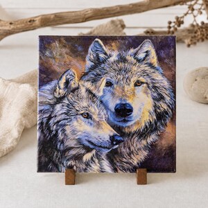 May include: Square tile featuring a detailed painting of two wolves. The artwork showcases the wolves' fur in shades of gray, blue, and yellow, set against a warm, brown background. The tile is displayed on a small wooden stand.