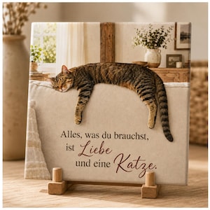 Könnte beinhalten: Ein rechteckiger Leinwanddruck mit einer braunen getigerten Katze, die auf einer beigen Oberfläche ruht. Der Text lautet: Alles, was du brauchst, ist Liebe und eine Katze. Der Druck wird von zwei kleinen Holzblöcken an der Basis gestützt und zeigt warme, neutrale Farben.