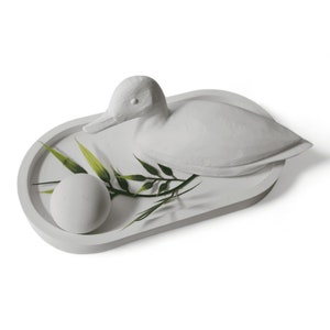 May include: A white ceramic tray with a white ceramic duck and a small white sphere. Green leaves are arranged around the sphere.