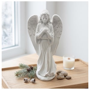 May include: A white angel statue with outstretched wings, standing on a wooden tray. A lit candle in a glass jar, small stones, and a sprig of pine with pinecones are also on the tray. The angel is the focal point of this decorative display.