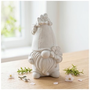 May include: A white garden gnome statue with a long white beard, a large nose, and a pointed hat adorned with flowers. The gnome is standing on a wooden surface, with sprigs of greenery and flowers scattered around it.
