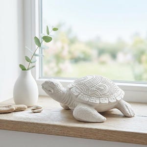 Könnte beinhalten: Eine weiße Keramik-Schildkrötenfigur mit detaillierten Schalenmustern steht auf einer Holzoberfläche. Eine kleine weiße Vase mit grünen Blättern und eine Muschel ergänzen das Küstenmotiv. Die Schildkröte ist ein dekoratives Wohnaccessoire.