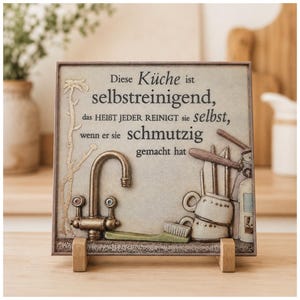 Könnte beinhalten: Eine quadratische Keramik-Wandtafel mit einem Relief-Design aus einem Küchenwasserhahn, einer Scheuerbürste und gestapelten Tassen. Der Text lautet: Diese Küche ist selbstreinigend, das HEIßT JEDER REINIGT sie selbst, wenn er sie schmutzig gemacht hat.