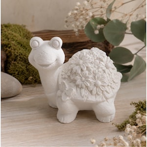 Op de afbeelding: Een witte keramische schildpadfiguur met een lachend gezicht en een schild versierd met bloemendetails. De schildpad heeft vier korte poten en staat tegen een natuurlijke achtergrond met groen en een houten element.