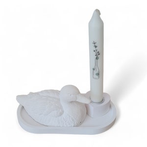 Op de afbeelding: Witte keramische kaarshouder in de vorm van een eend met een witte kaars en een bloemenmotief. De kaarshouder staat op een witte basis.